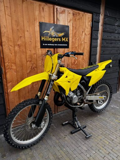 Geel crossmotor staat naast een houten muur met een logo erop.